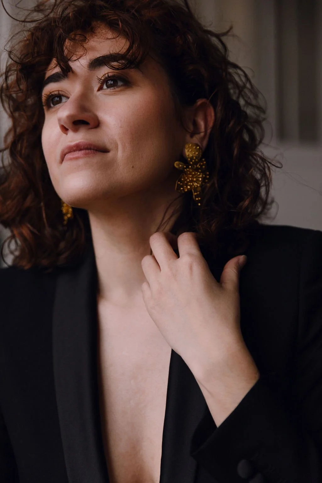 Close-up of Alex Vidal with curly brown hair, wearing gold earrings and a black blazer, looking away with a thoughtful expression.