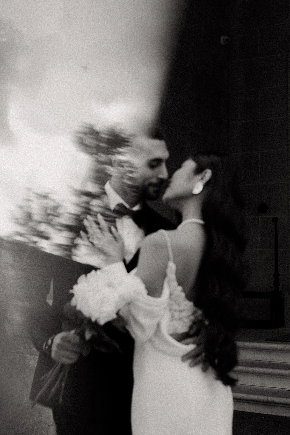 A couple sharing a dance indoors, with the man wearing a tuxedo and the woman in a white dress holding a bouquet of white flowers, blurred due to motion.