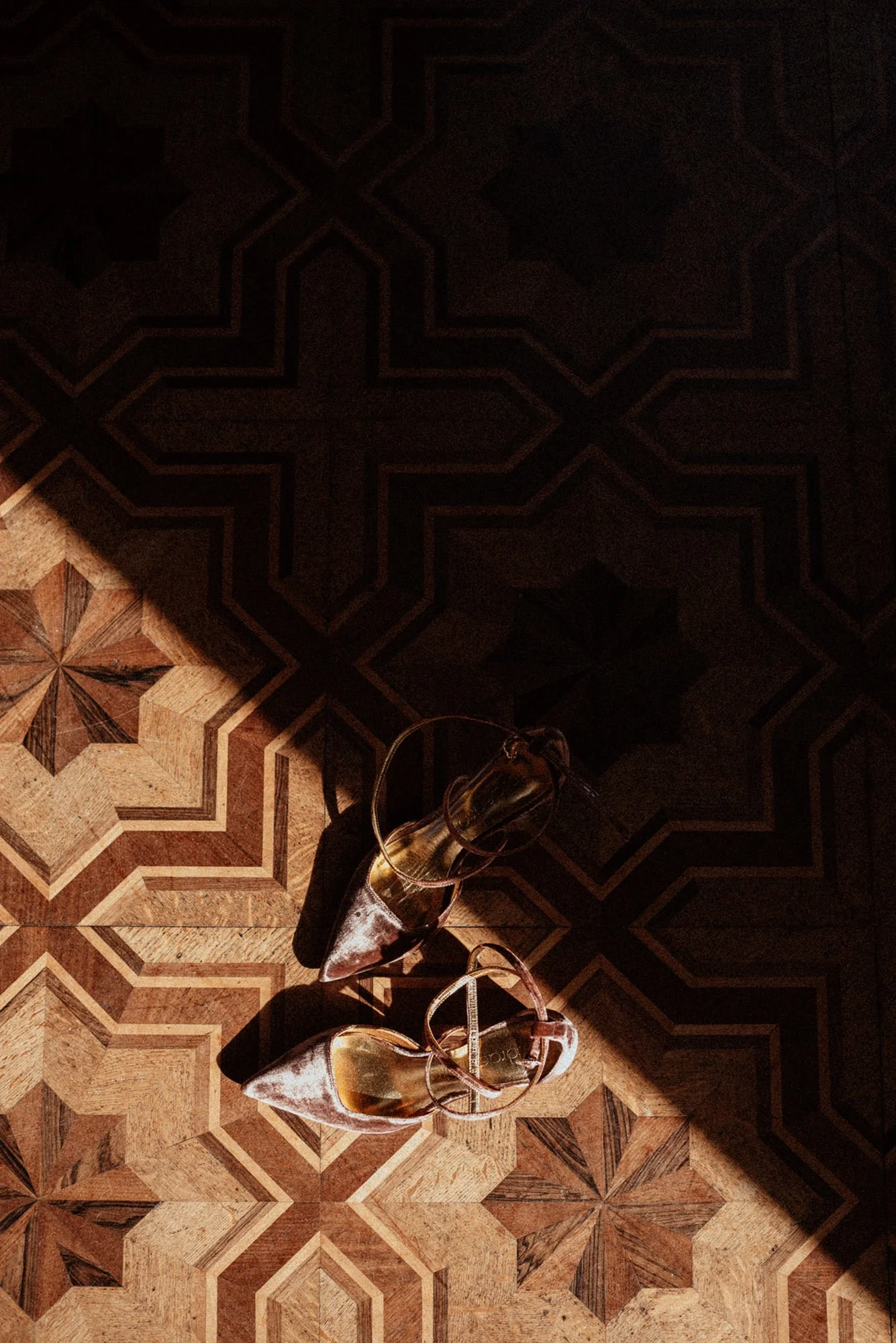 A close-up of a pair of burgundy velvet bridal shoes, resting on a beautiful inlaid wooden floor, illuminated by a beam of light, in a chiaroscuro photograph