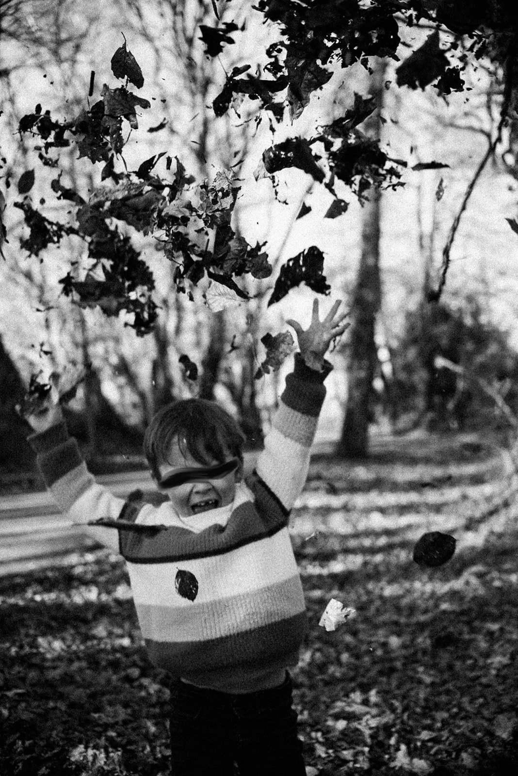 black-and-white photo of a little boy tossing a pile of autumn leaves into the air, with a missing front tooth