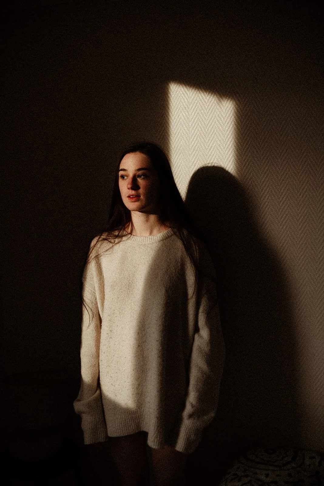 A young woman with long dark hair standing against a wall, illuminated by a rectangular patch of light, wearing a cream-colored sweater.