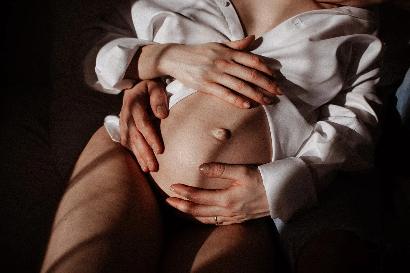 Close-up of two people holding a pregnant belly, wearing a white shirt, with dark background.