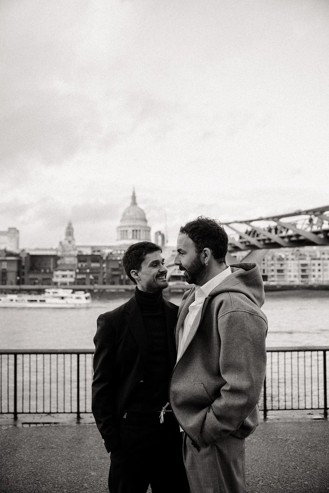 Two men smiling and facing each other outside near the Tames with London cityscape in the background, black and white photo.