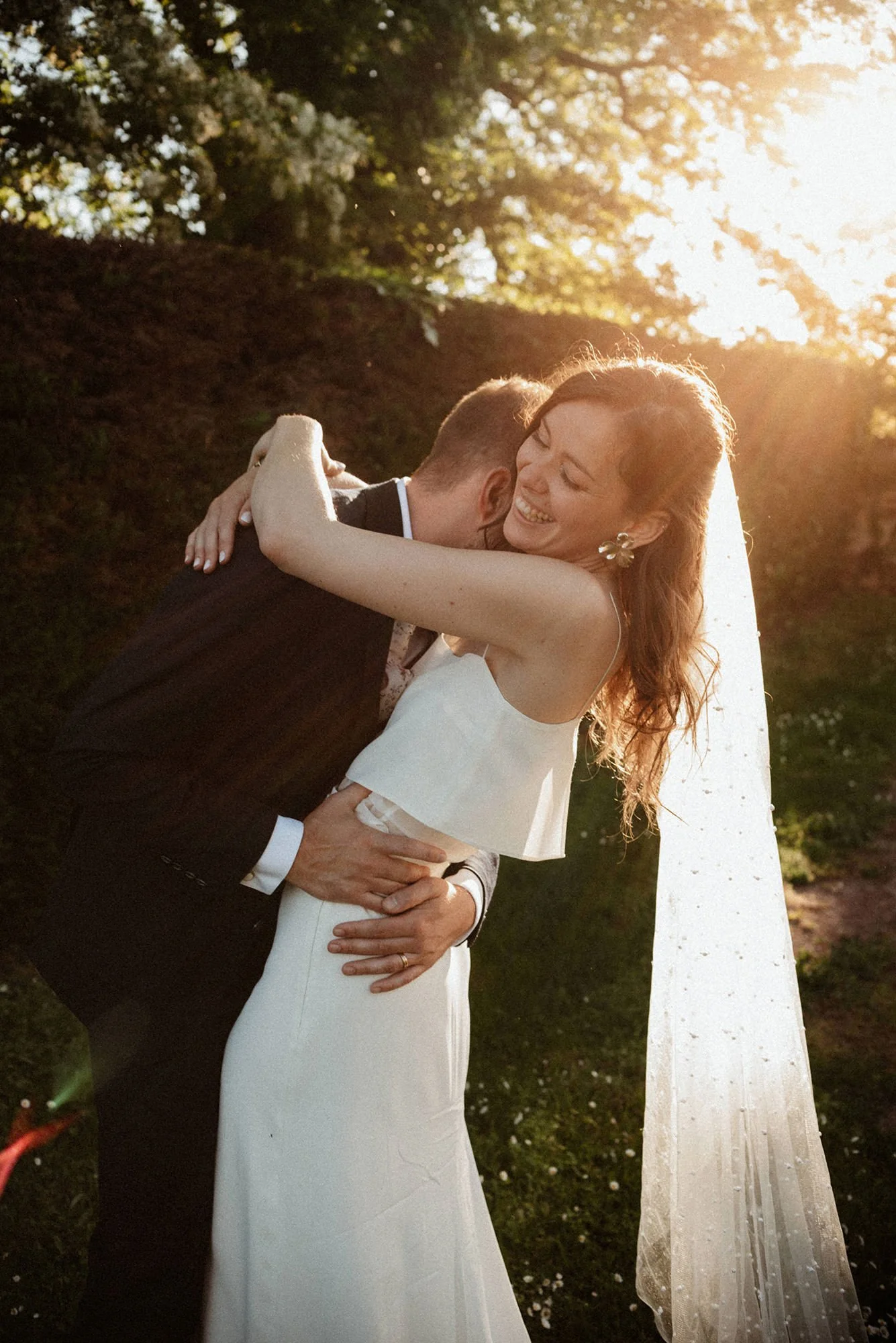 a newlywed couple at sunset, bathed in a halo of light, the groom’s face hidden in his wife’s neck as she laughs heartily, her arms wrapped around his neck