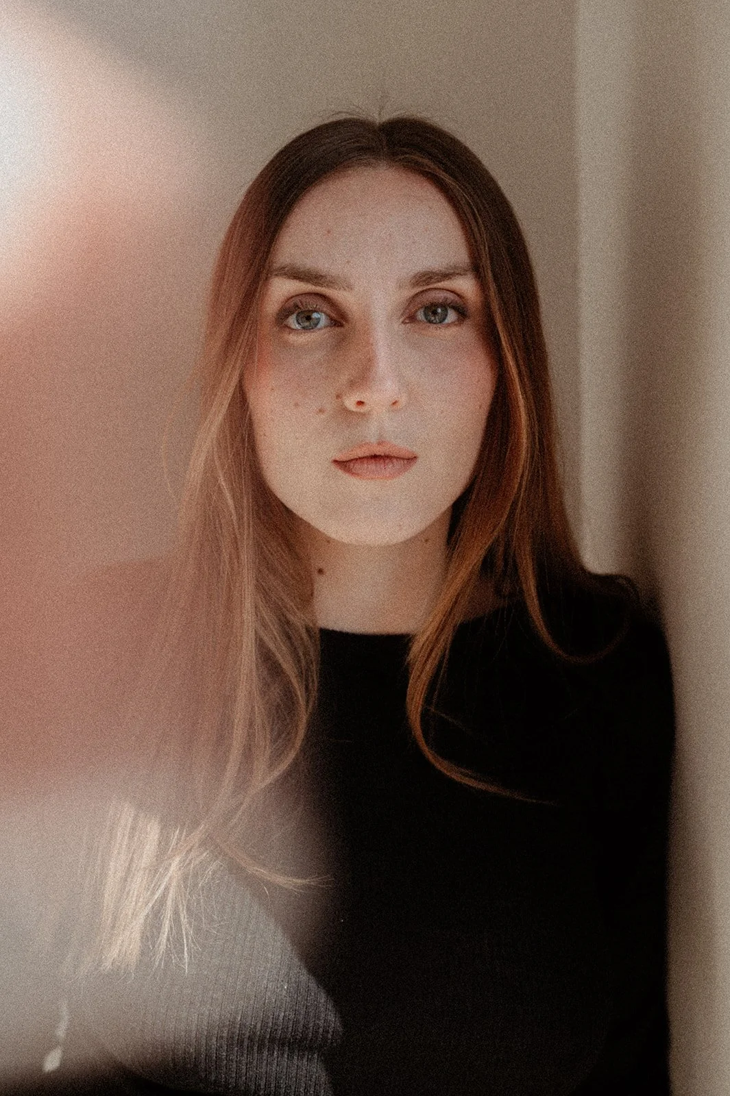 Portrait of a young woman with light skin, long brown hair, and blue eyes, wearing a black top, standing against a plain wall.