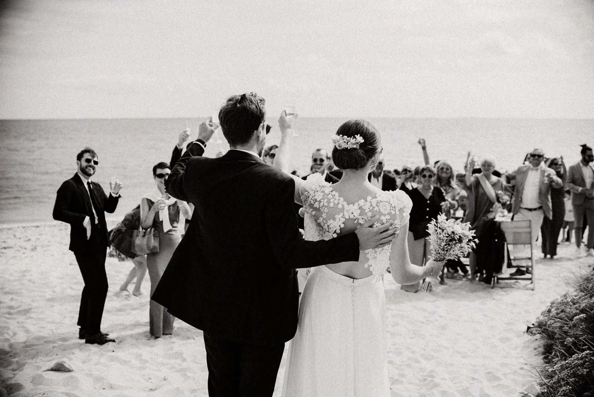 A black-and-white photo of a couple, seen from behind, standing in front of their guests on the beach, celebrating their wedding by raising a glass of champagne, with the groom’s hand resting on the bride’s back