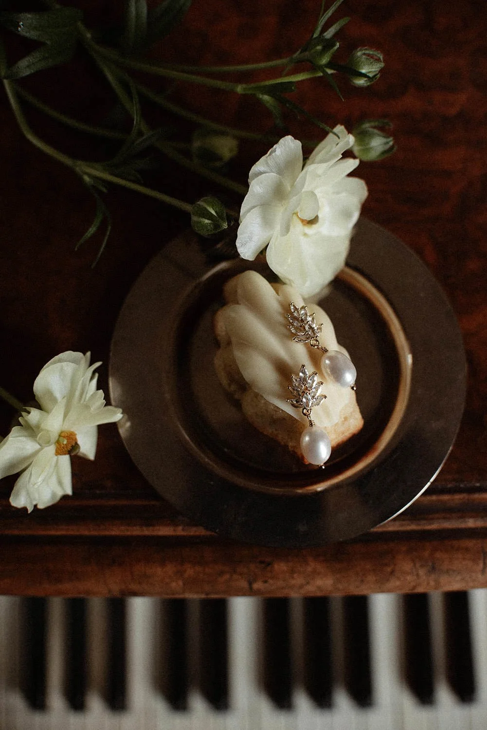 A jewelry ring with pearl drops and leaf-shaped gemstones resting on a shell, surrounded by white flowers in a small black dish on a wooden surface.