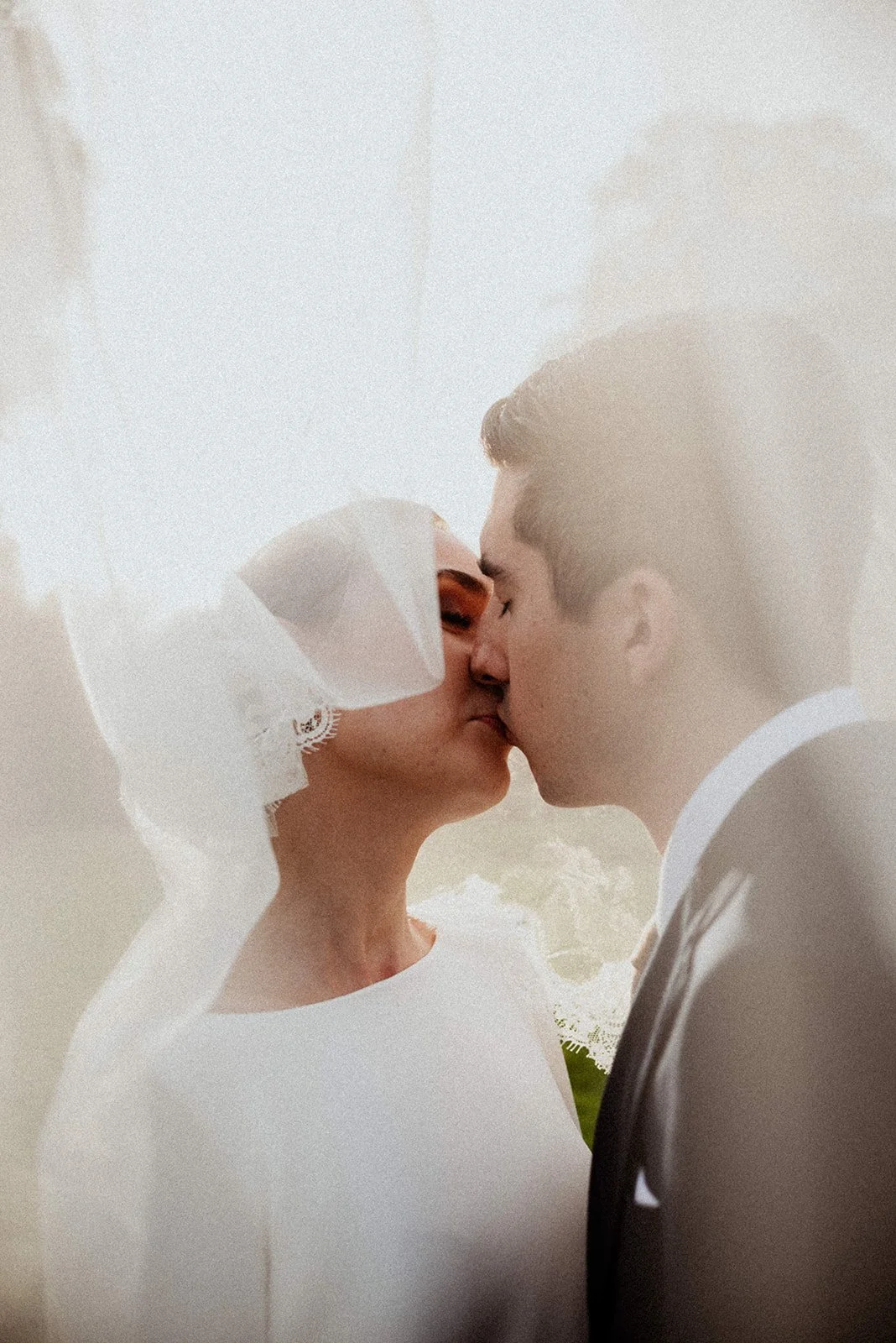 A bride and groom share a kiss, with the bride wearing a white dress and lacy veil, and the groom in a suit and tie, outdoors with a bright background.