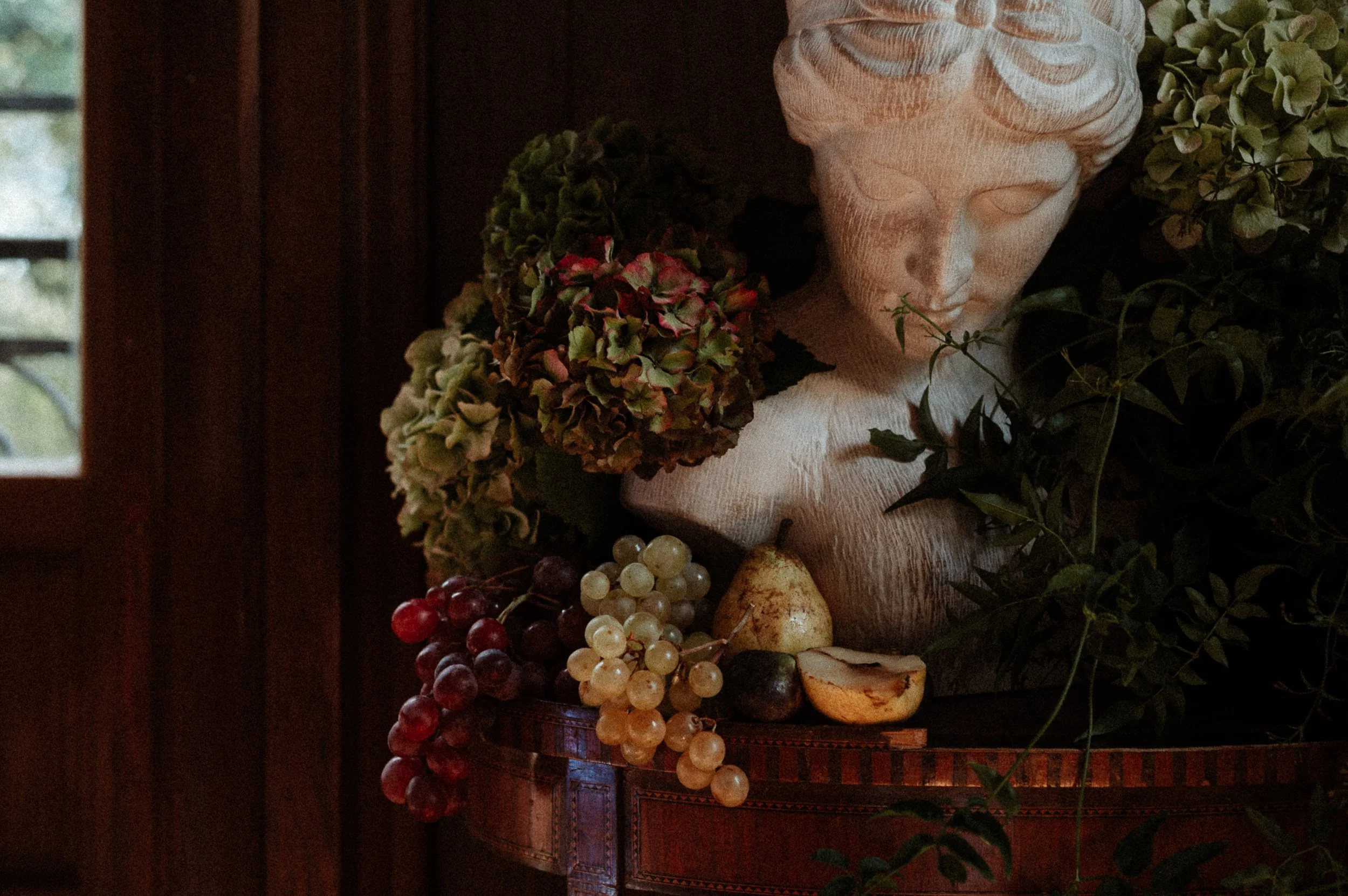 A decorative sculpture of a woman's face surrounded by dried hydrangea flowers, grapes, two pears, and leafy greenery on a wooden table in a dimly lit room.