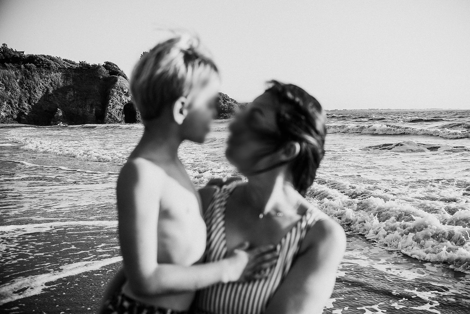 A black and white photo of a mother and her boy, close to the shoreline with waves, rocks, and cliffs in the background, embracing and looking at each other.