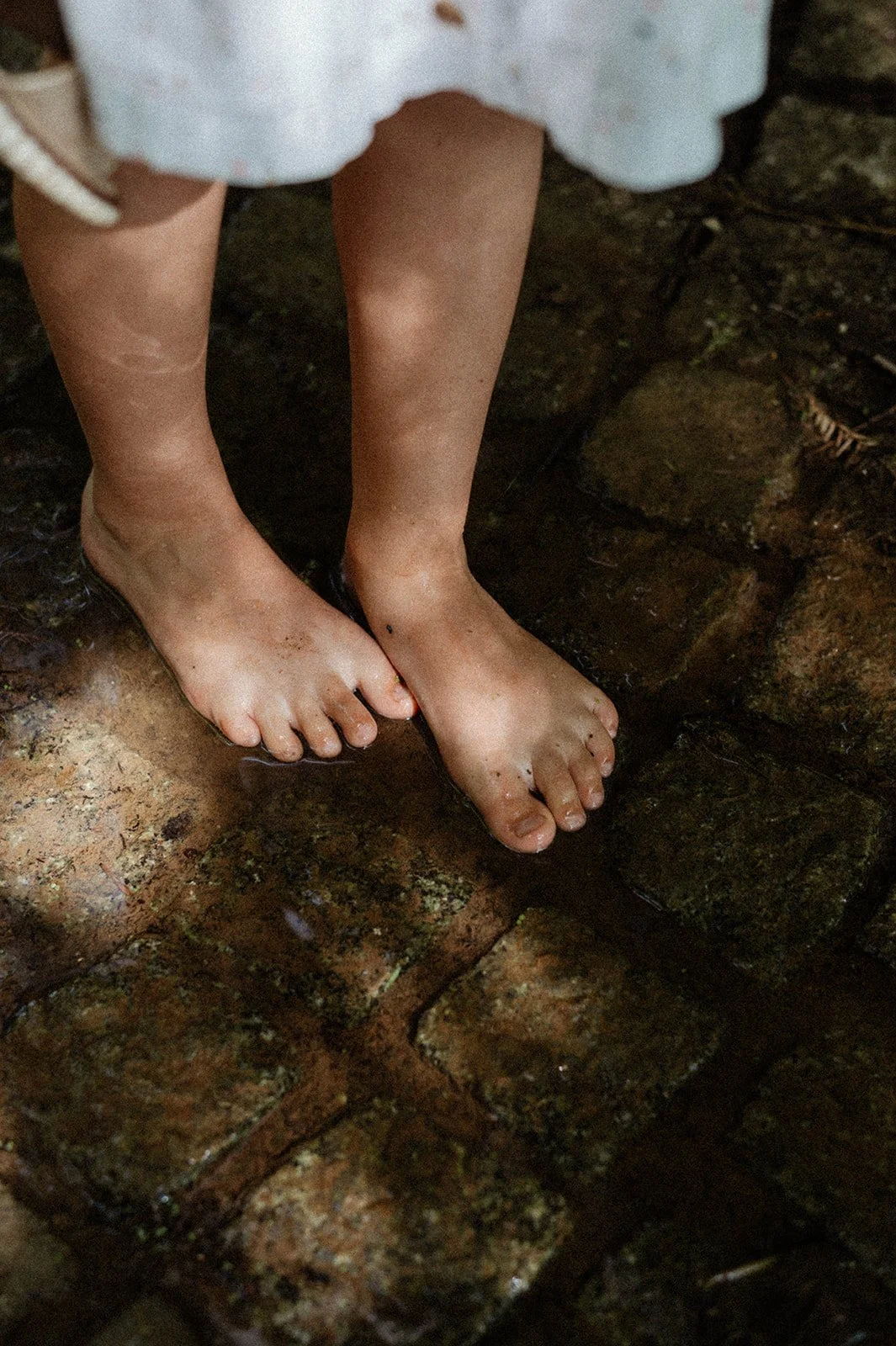 Child's bare feet on wet brick stones, with part of a white dress visible.