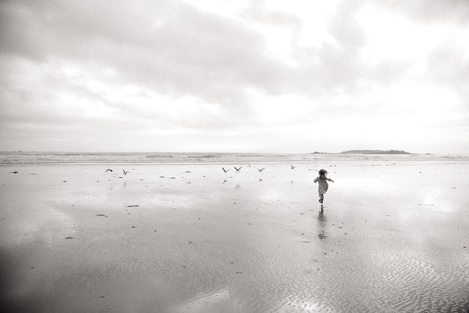 A black and white photo of a child running on a beach with seagulls flying and a distant landmass on the horizon.