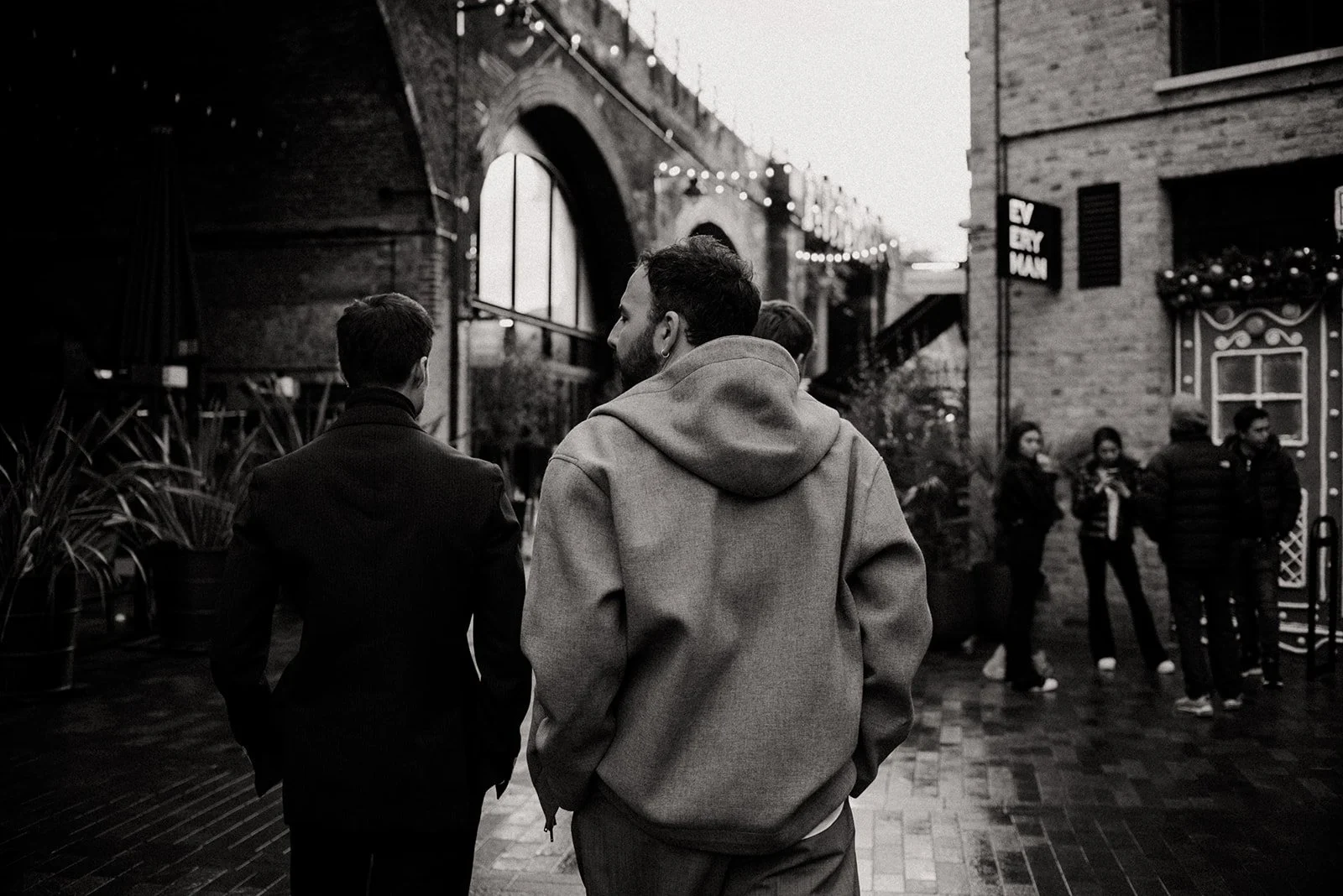 People walking in an urban street of London, including a couple of two men in the foreground, with Christmas lights and storefronts visible.