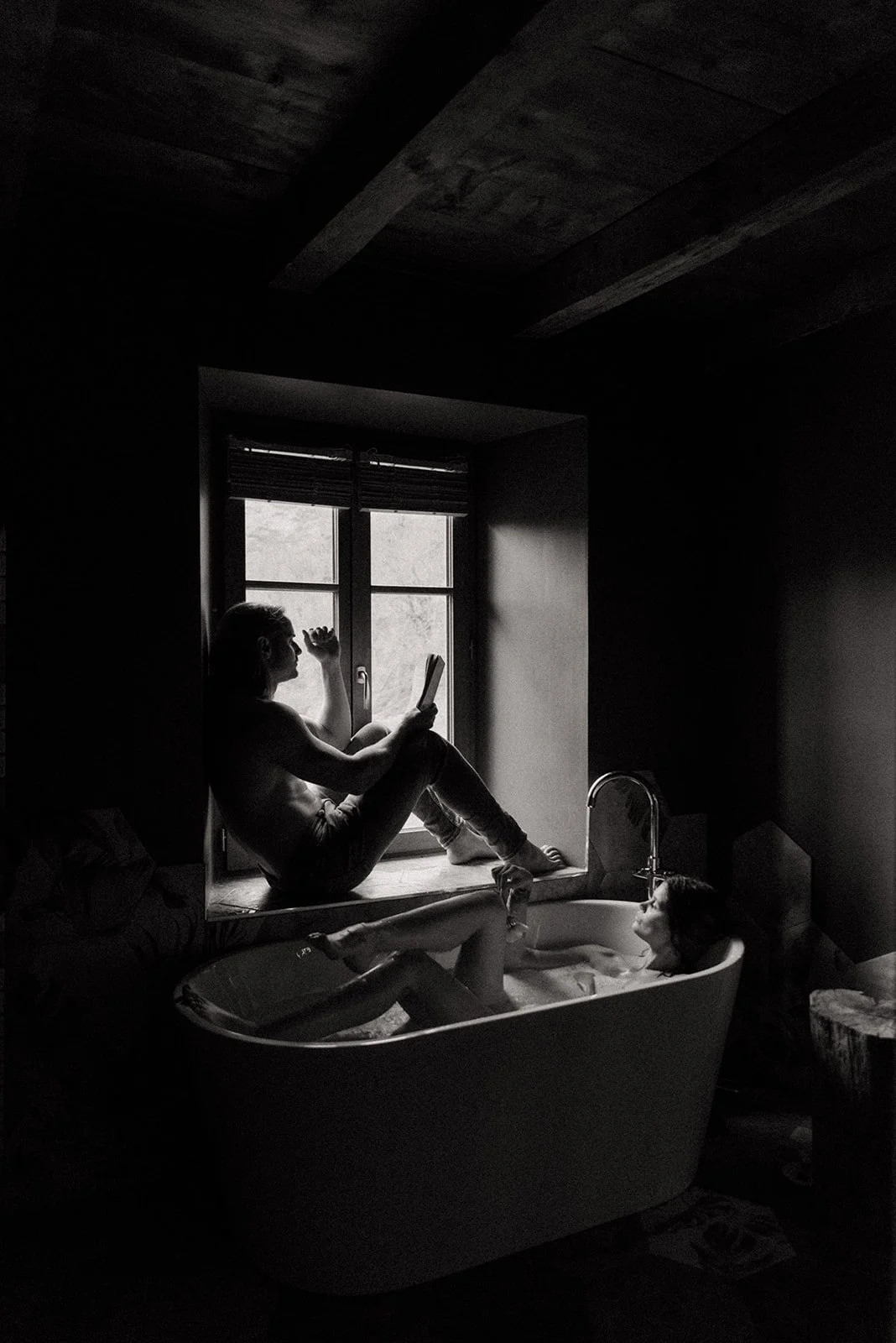 A black and white photo of a woman sitting on a window ledge reading a book, with her partner lying in a bathtub nearby in a dimly lit room with a wooden ceiling.