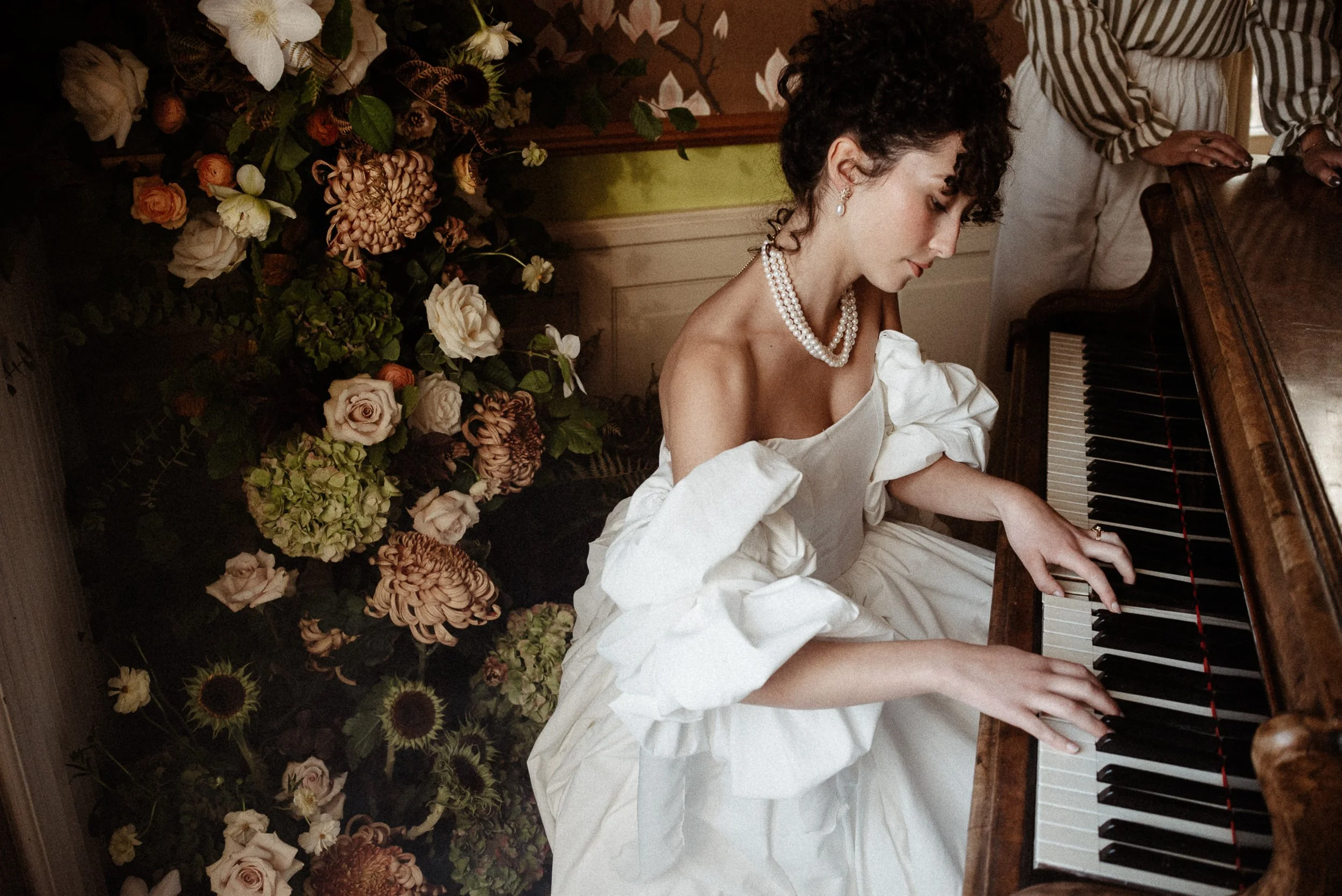 A woman dressed in a vintage white wedding dress, wearing pearl jewelry, is playing the piano. She is sitting in front of a wall decorated with an abundance of colorful flowers and greenery.