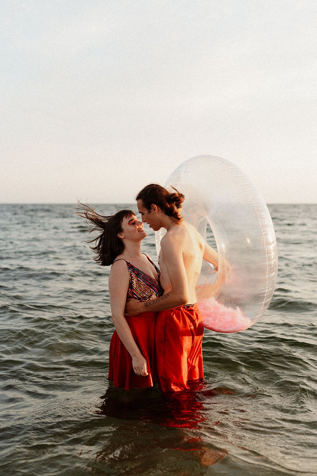 wide shot of a couple dressed in multicoloured sequins, embracing and submerged up to their thighs in the sea; his hand is on her waist, her hair blowing in the wind; he is holding a huge transparent ring float with pink feathers