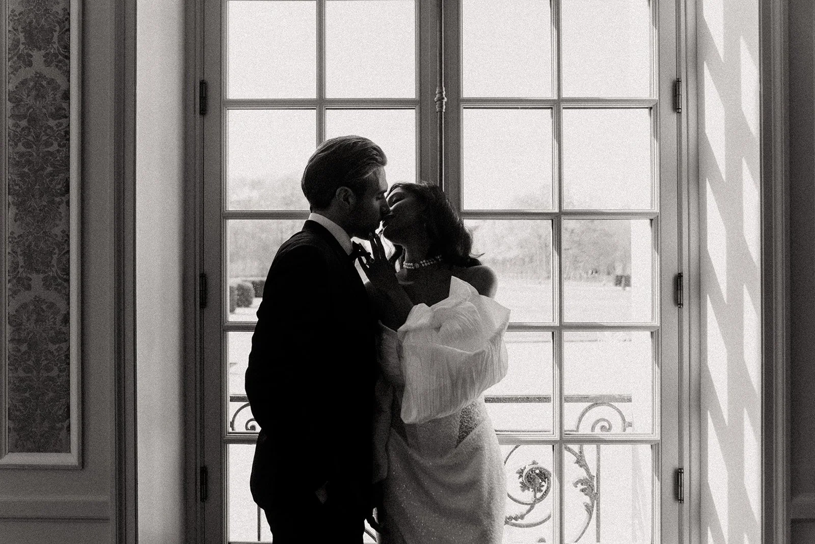 A black and white photo of a couple kissing in front of a large window, with sunlight creating shadows on the window and floor.