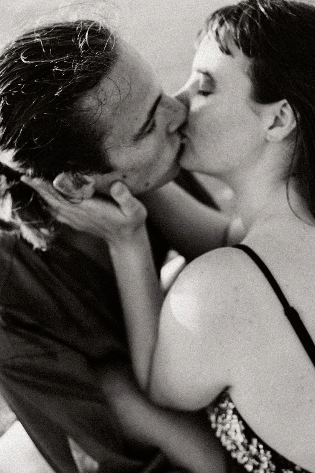 A close-up shot from above of a couple embracing and kissing, the woman’s hand tenderly caressing her partner’s cheek; she is wearing sequins; black-and-white photograph
