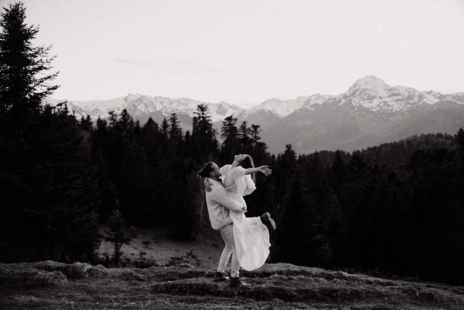 A couple dressed in wedding attire dancing in a forested mountainous landscape with snow-capped mountains in the background, depicted in black and white.