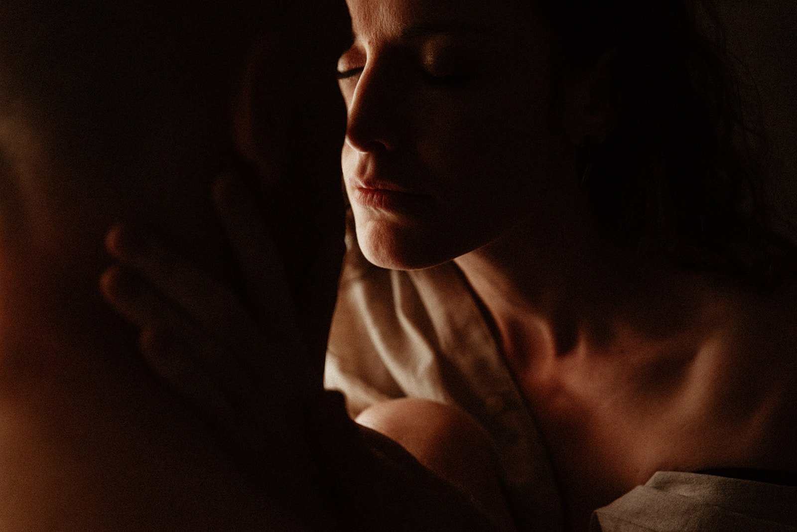 A close-up of a woman’s face, her eyes closed, as she leans in to kiss her partner, her hands cupping his face, in chiaroscuro, soft lighting
