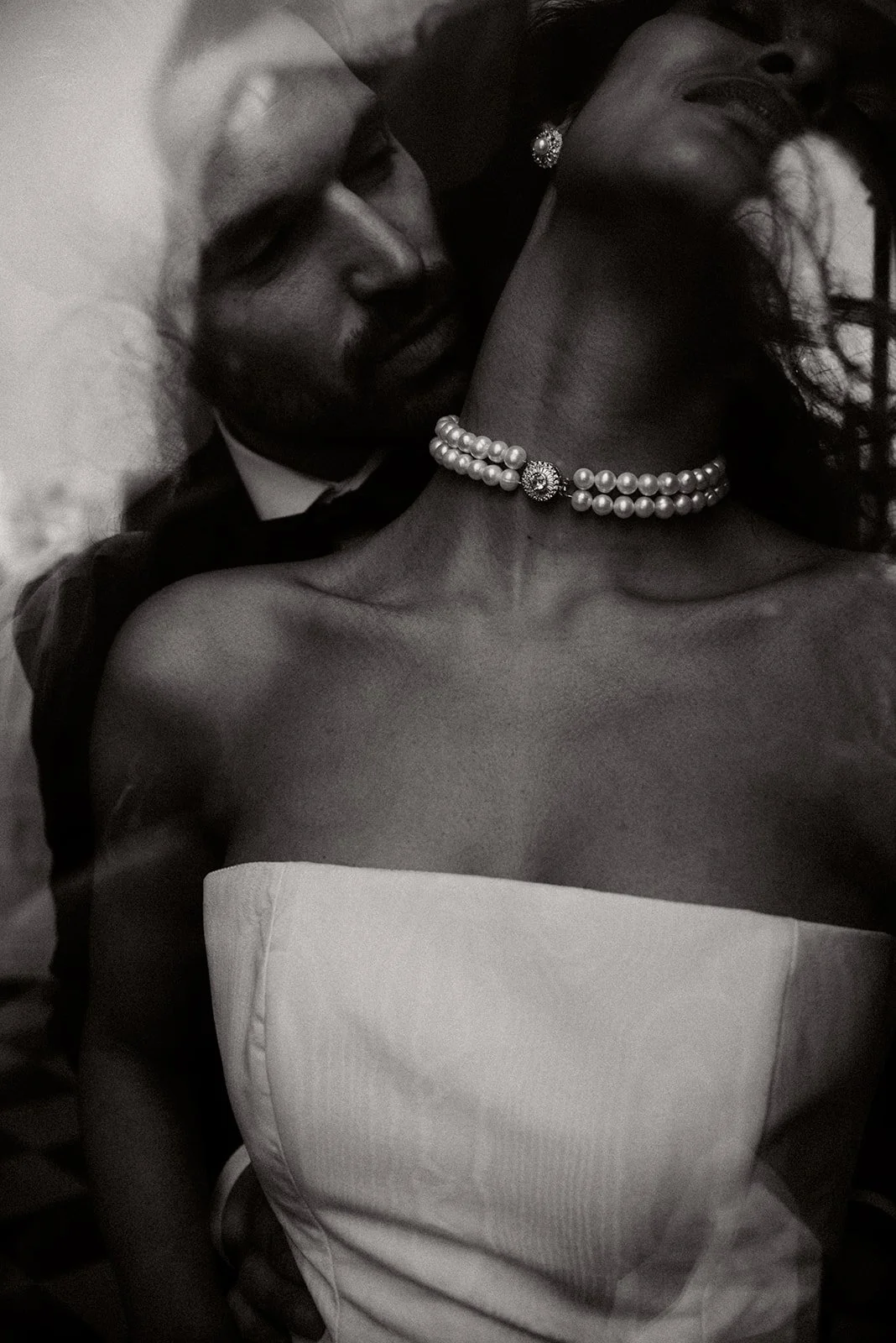 A close-up black and white photo of a man and woman in an intimate pose. The man is leaning close to the woman's neck, smiling, with a beard and wearing a suit. The woman is wearing a strapless dress, a pearl necklace, and earrings, with long wavy hair.