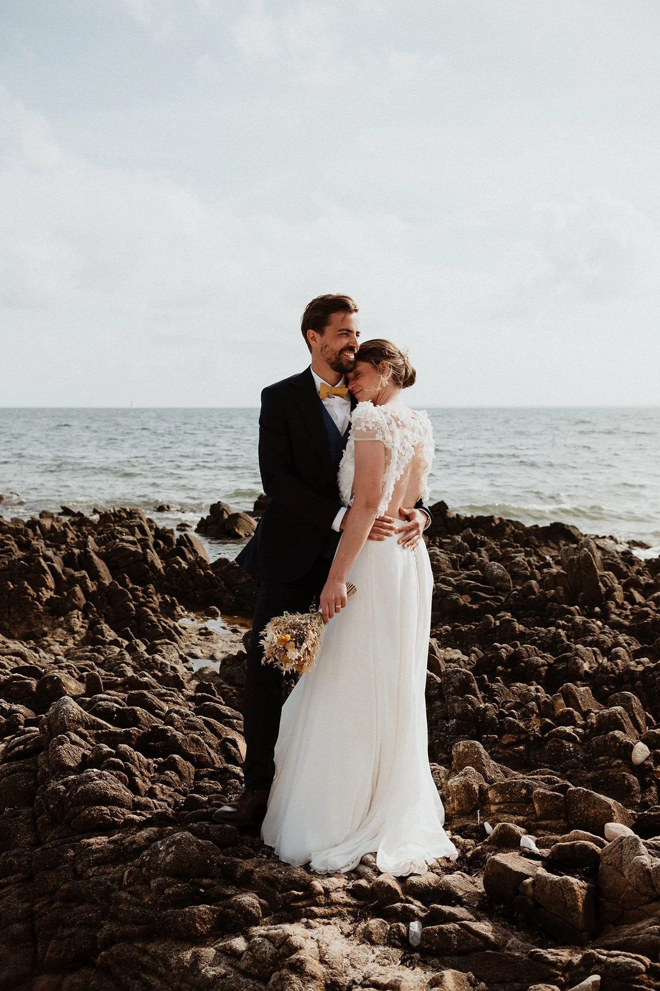 the bride and groom embracing, standing on the rocks by the sea, the groom’s hands resting on his wife’s back, her bare back revealed by her lace dress; they are smiling
