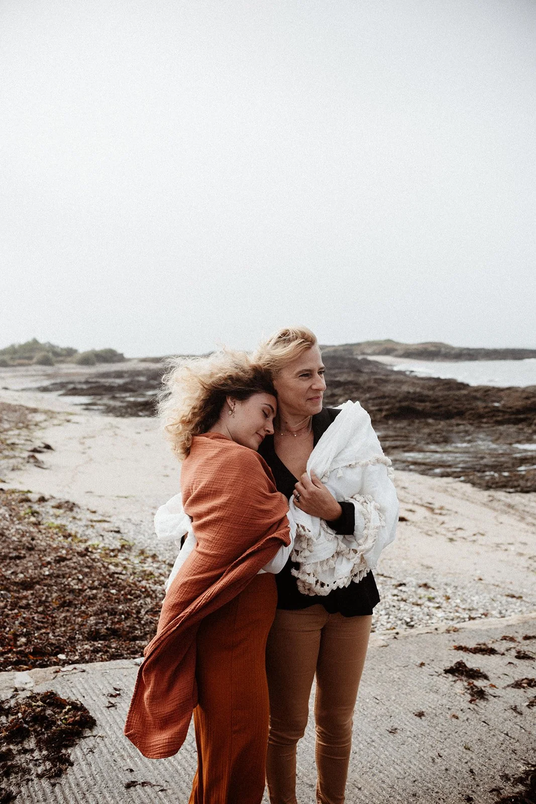 Two women hugging on a beach, one with curly hair and a rust-colored outfit, the other with straight blonde hair holding a white blanket, looking thoughtfully into the distance.