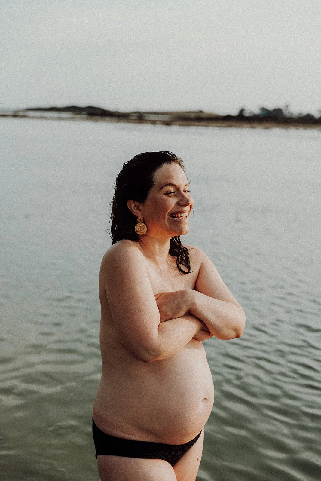 A woman with dark hair and gold earrings smiling with her arms crossed, standing in water at a beach during daylight.