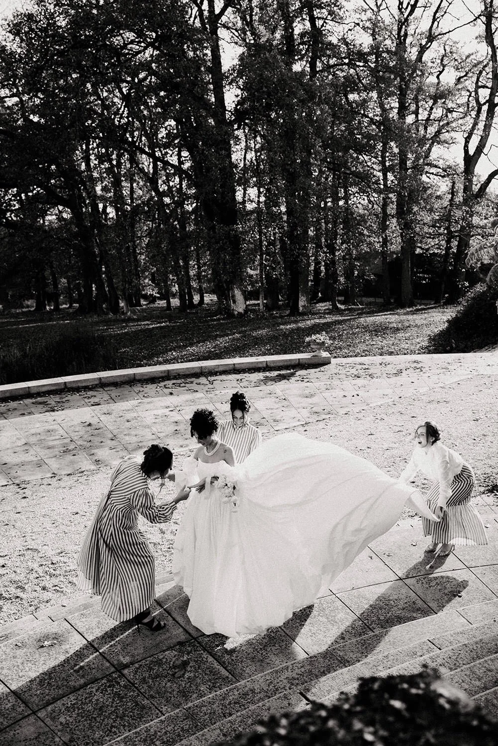A black-and-white overhead shot of a bride surrounded by her three bridesmaids, who are admiring her wedding ring and lifting her long train into the air to spread it out on the ground, on the terrace of a château
