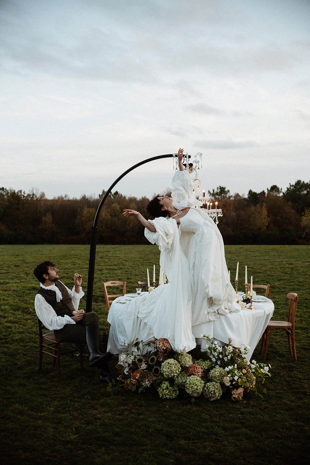 A couple dressed in wedding attire is seated and standing around a long, decorated table outdoors, with the bride dancing on the table, all under a large, curved metal arch with candles and floral arrangements.