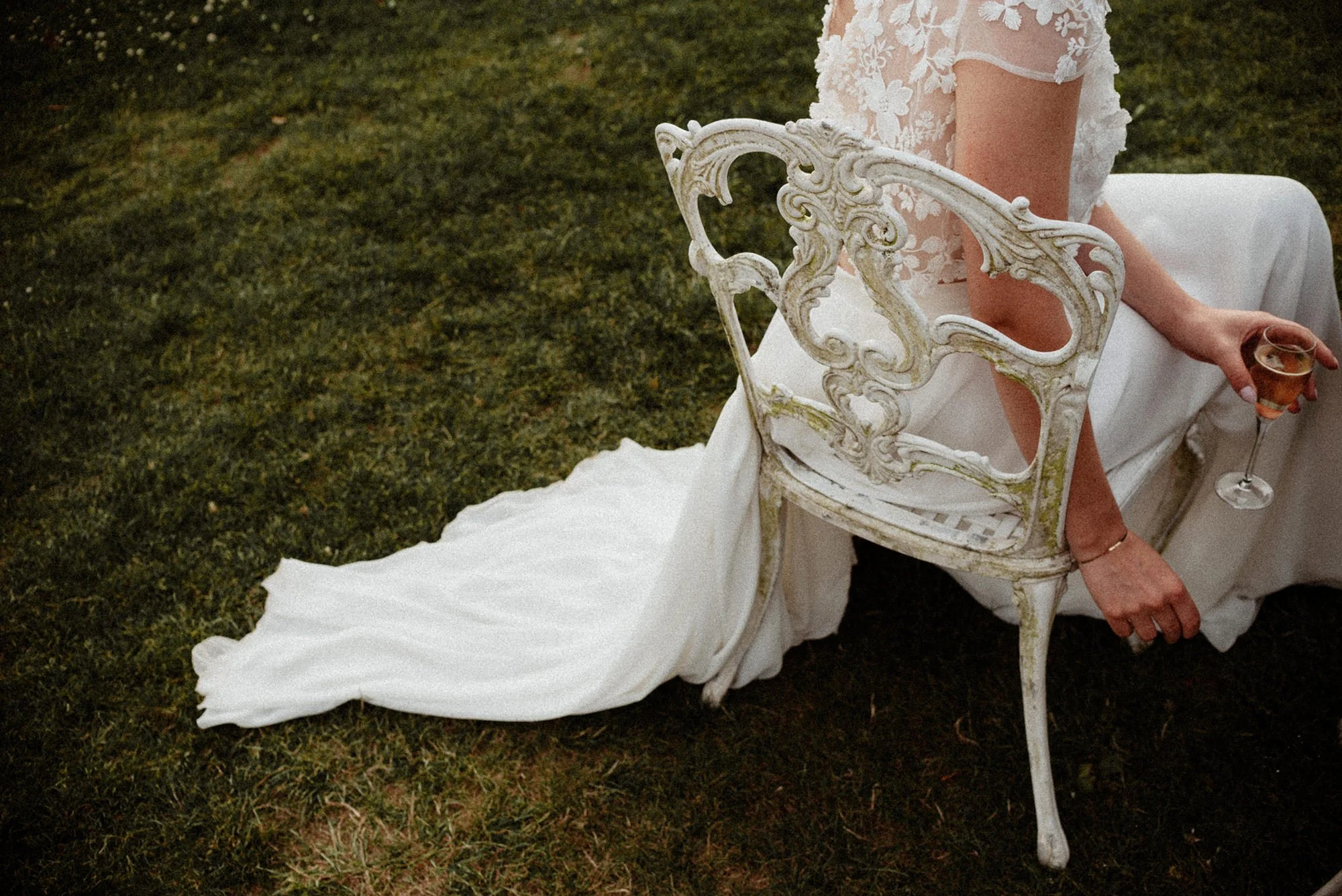 A close-up of a bride’s dress, seen from behind, as she sits on a beautiful white cast-iron chair in the garden during cocktail hour; her train contrasts with the green of the lawn, and she is holding a champagne flute in her hand