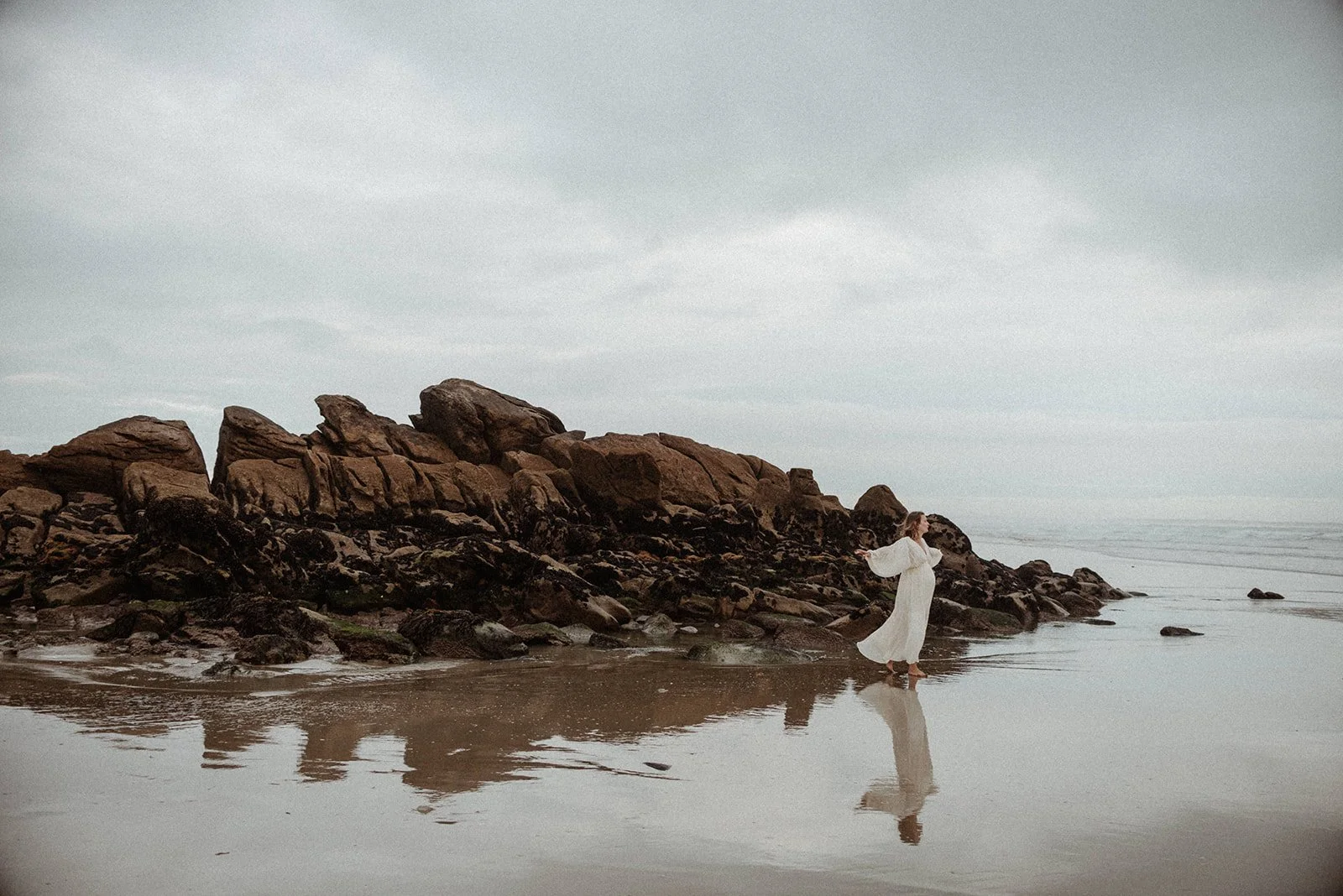 A wide shot of a woman wearing a long ivory dress that flutters in the wind, dancing with her arms outstretched on a misty beach, with a cluster of dark rocks behind her