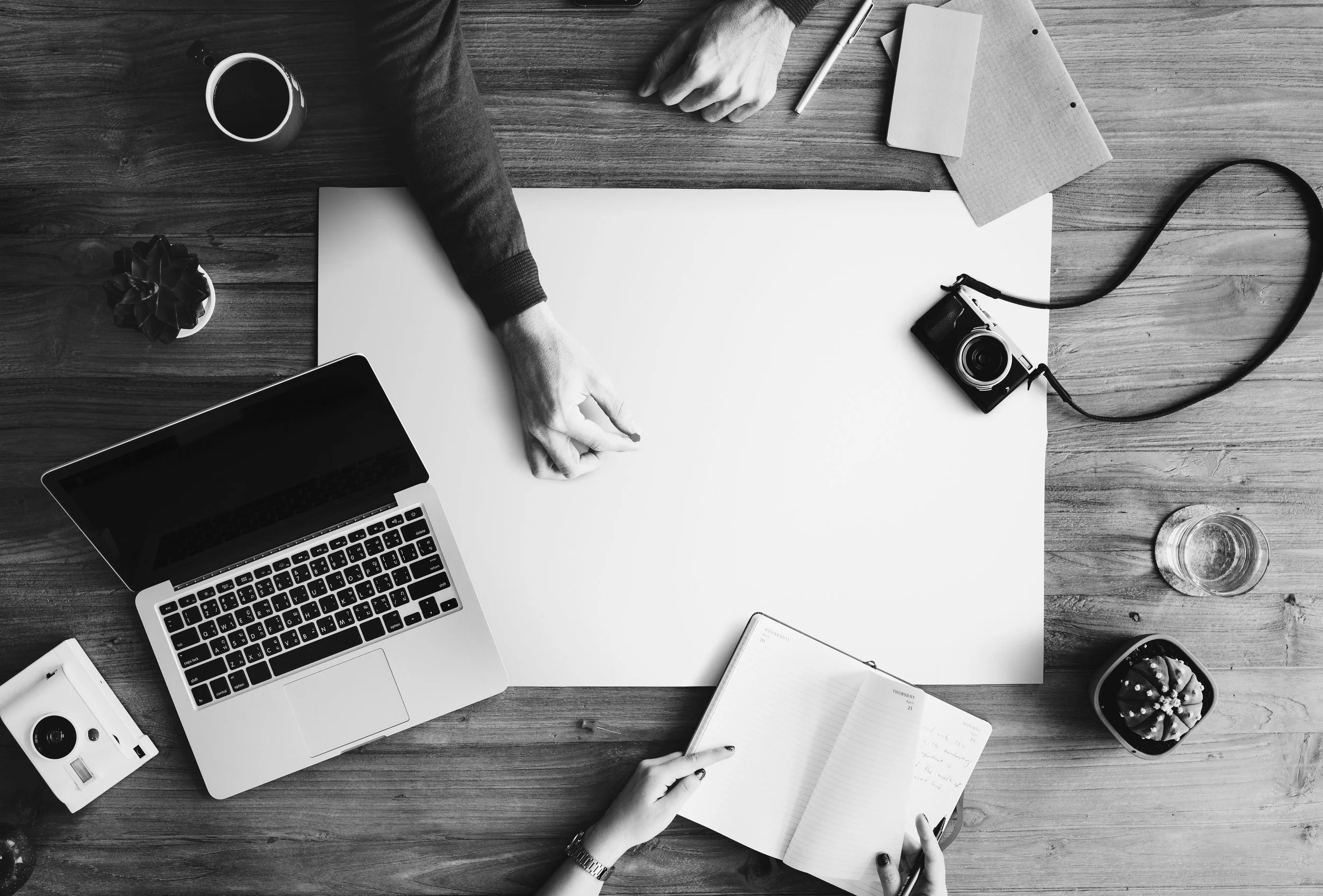 Top-down view of a workspace with a laptop, camera, notebooks, a notebook, a cup of coffee, a glass of water, a small plant, and hands of two people, one pointing to a notebook and the other holding a pen, on a wooden table.