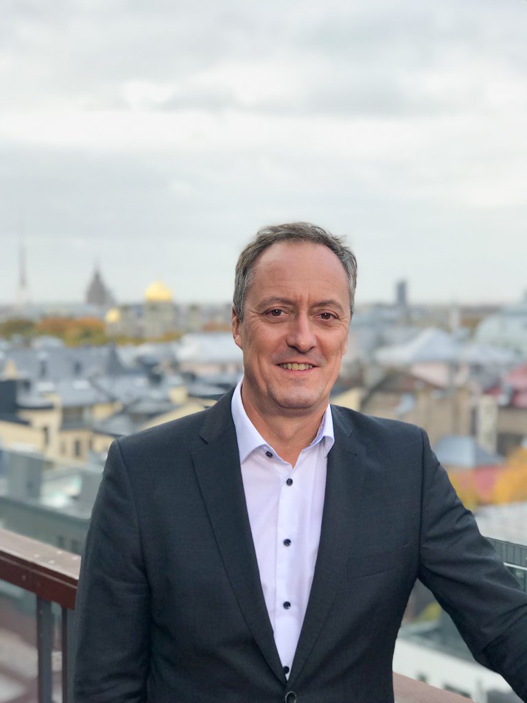 Ein Mann in einem schwarzen Anzug und weißem Hemd steht auf einem Balkon mit Blick auf eine Stadt mit Hindergrund. Es ist bewölkt.