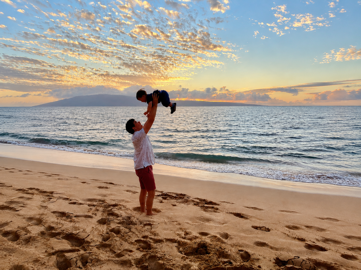 A man lifting a child on a beach at sunset with clouds and a mountain in the background.