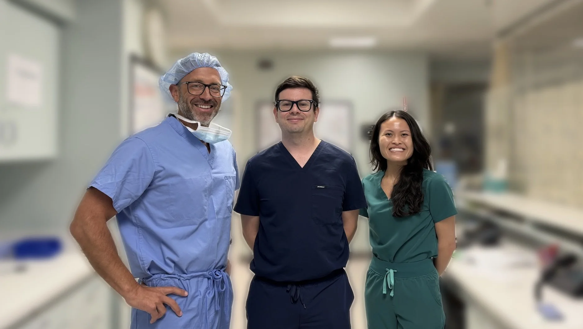 Three healthcare professionals standing in a medical facility, wearing scrubs and smiling at the camera.