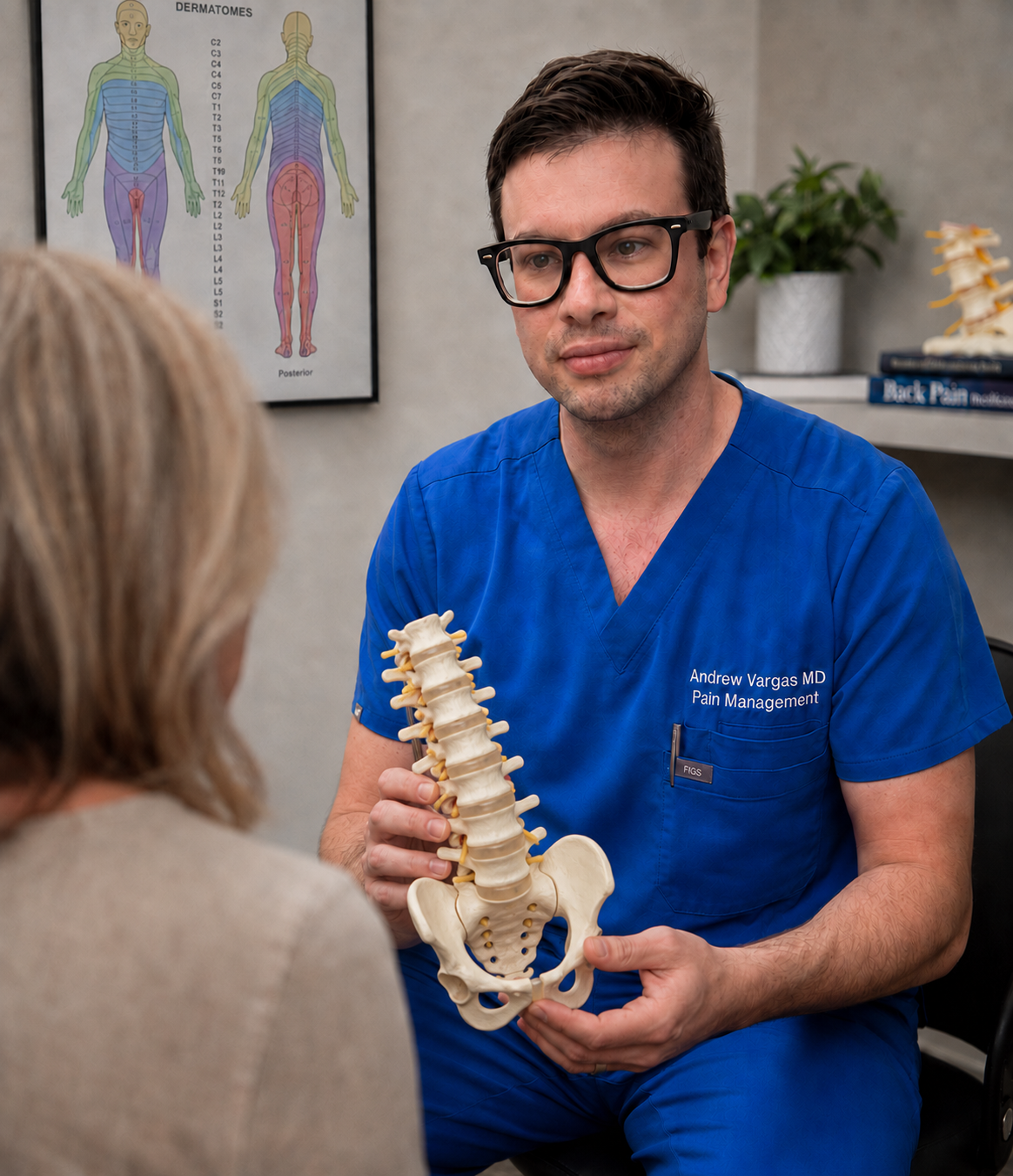 A male healthcare professional in blue scrubs showing a plastic model of a human spine to a woman patient, in a medical office setting with a dermatitis chart and spine model in the background.