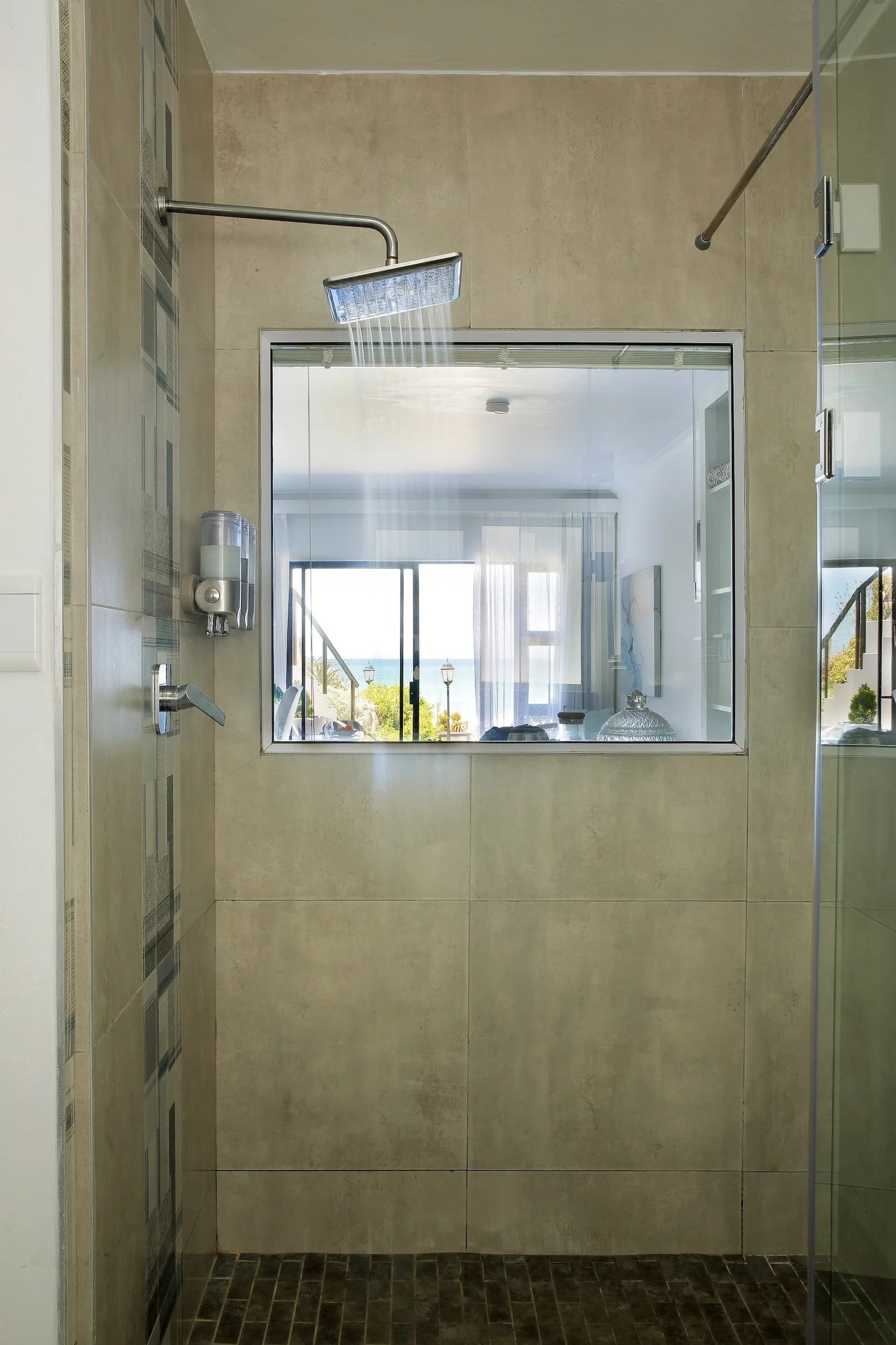 Modern en-suite bathroom with walk-in rain shower and feature window overlooking sea-facing bedroom in St Francis Bay guest house.