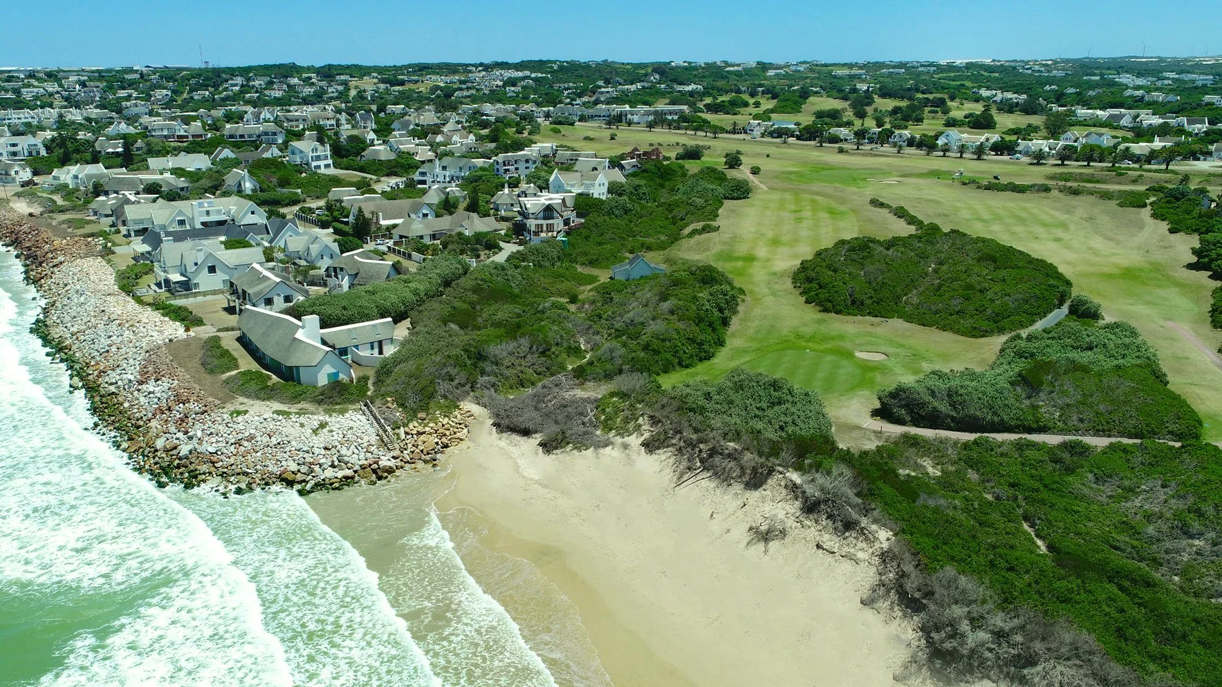 Aerial view of St Francis Bay coastline showing sandy beach, ocean waves, seaside homes, and a coastal golf course along the Eastern Cape shoreline, South Africa.