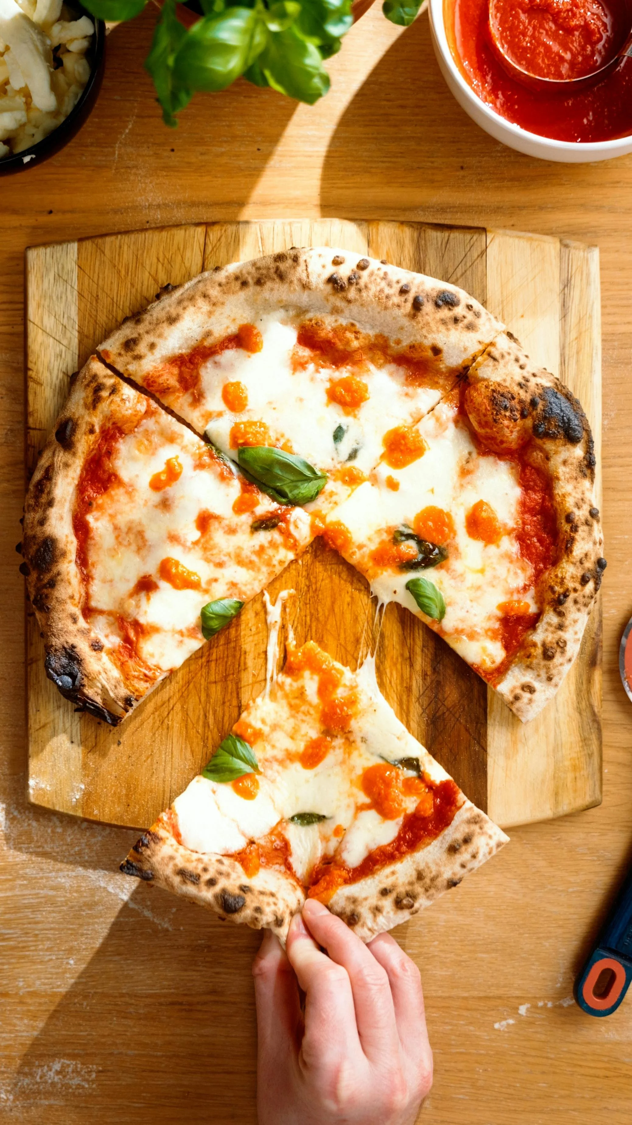 A pizza with a thin crust, topped with mozzarella cheese, basil leaves, and orange sauce, placed on a wooden cutting board. A hand is removing a slice of pizza.