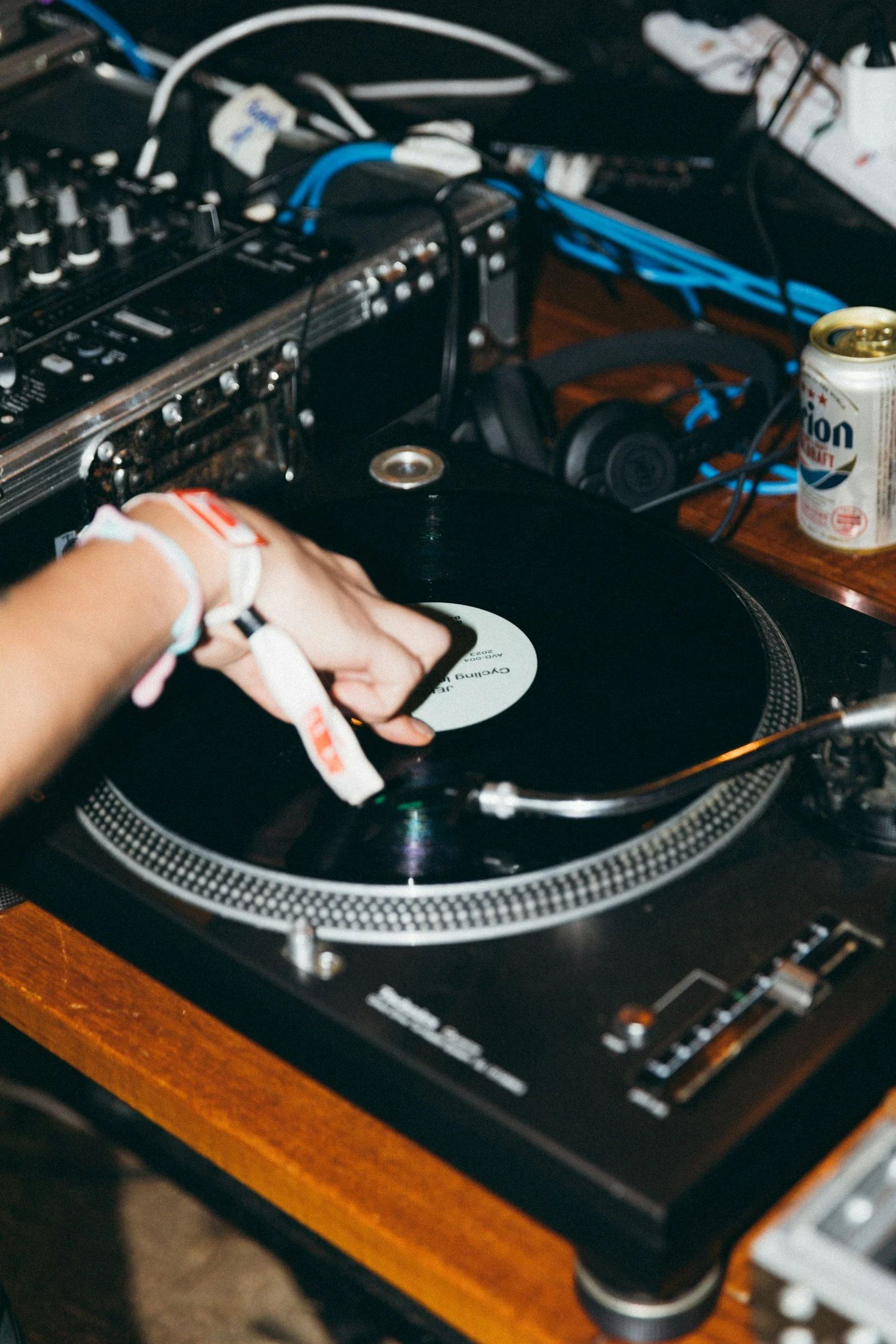 A person's hand on a vinyl record on a DJ turntable, with audio equipment, wires, a headphone, and a can of soda on a wooden table.