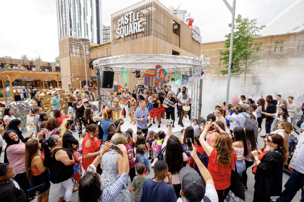 Crowd of people enjoying live music and dancing outdoors in an urban area with wooden structures and a building labeled 'Castle Square' in the background.