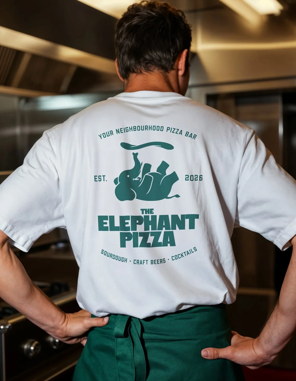 Person wearing a white T-shirt with a pizza promotional design, standing with hands on hips in a kitchen.
