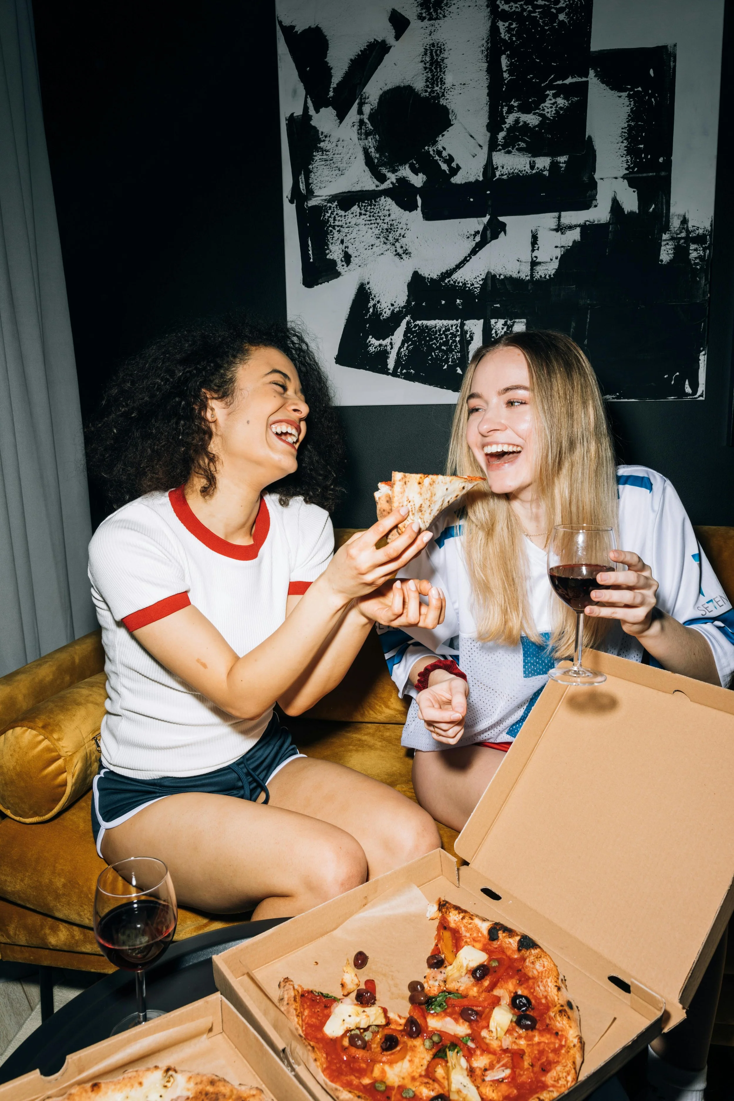 Two women sitting on a yellow couch, laughing and sharing a slice of pizza. One woman holds a glass of red wine, and an open pizza box with slices of pizza is in front of them. The background features black and white abstract artwork.