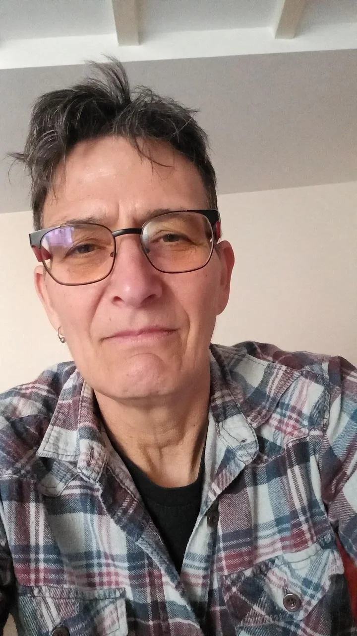 Un portrait d'une personne avec des lunettes, des cheveux en désordre, portant une chemise en flanelle à carreaux, prise en gros plan dans un intérieur.