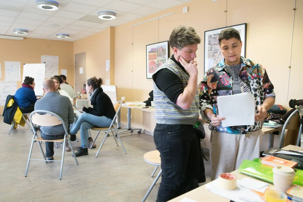 Deux femmes discutent et regardent un document dans un environnement de salle de réunion ou de formation, avec d'autres personnes en arrière-plan, dans une ambiance professionnelle.