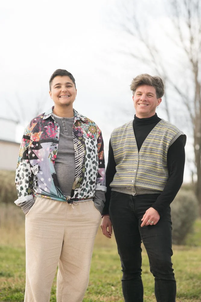 Deux personnes souriantes debout dans un parc, l'une portant un manteau multicolore avec des motifs, l'autre un pull à manches longues et un gilet, fond d'arbres sans feuilles.