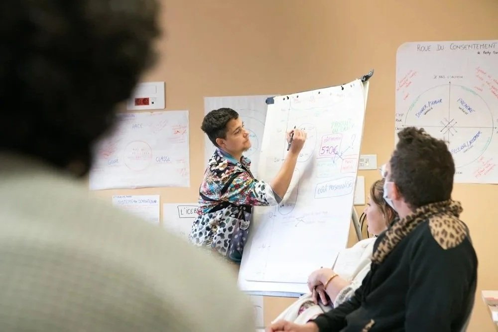 Un jeune homme présente lors d'une réunion ou d'un atelier, utilisant un tableau blanc pour écrire ou faire une présentation, entouré d'autres personnes qui écoutent attentivement.