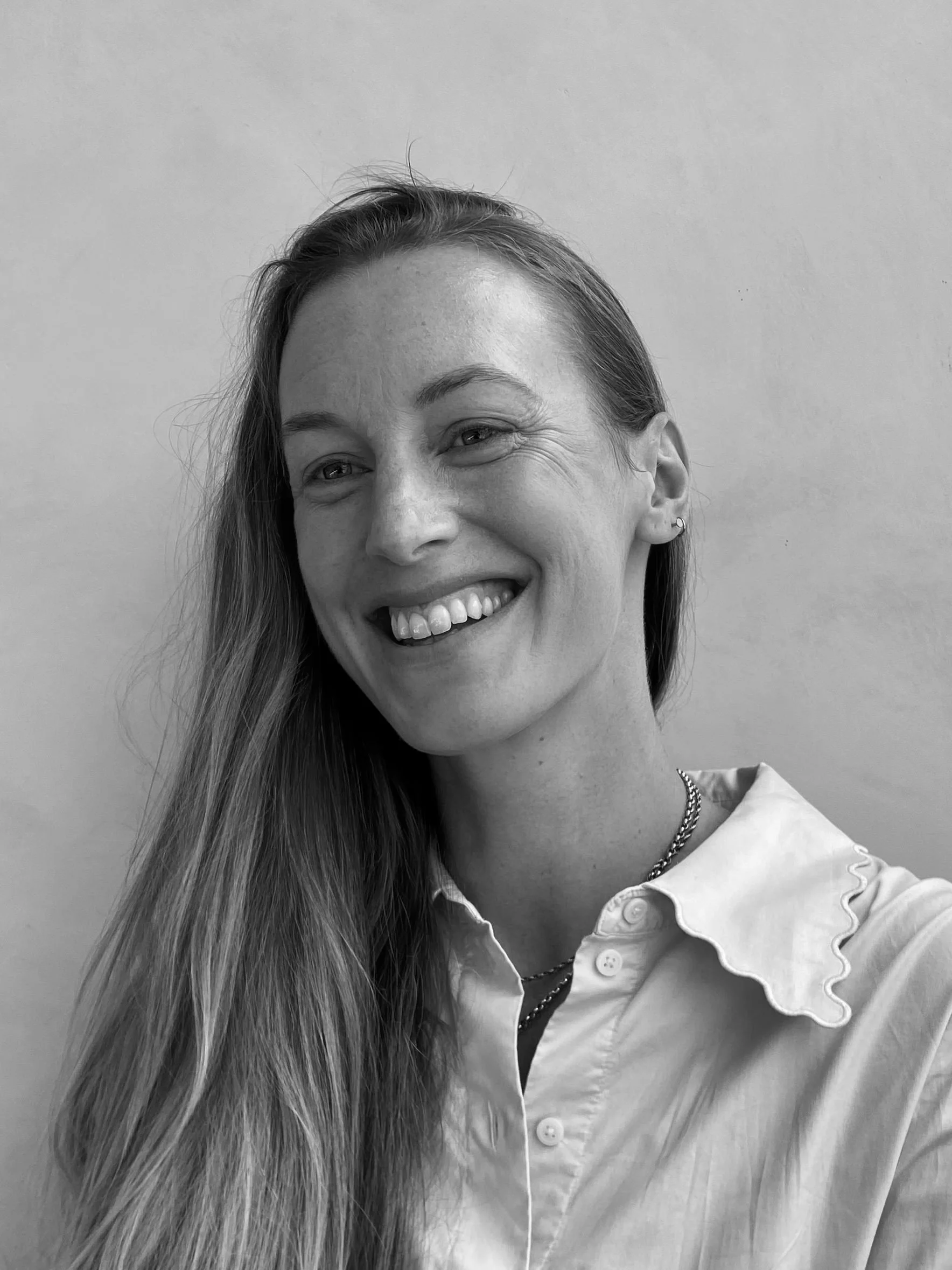 A grayscale photo of a woman smiling, showing her teeth, with long straight hair, wearing a collared shirt and a necklace, standing against a plain background.