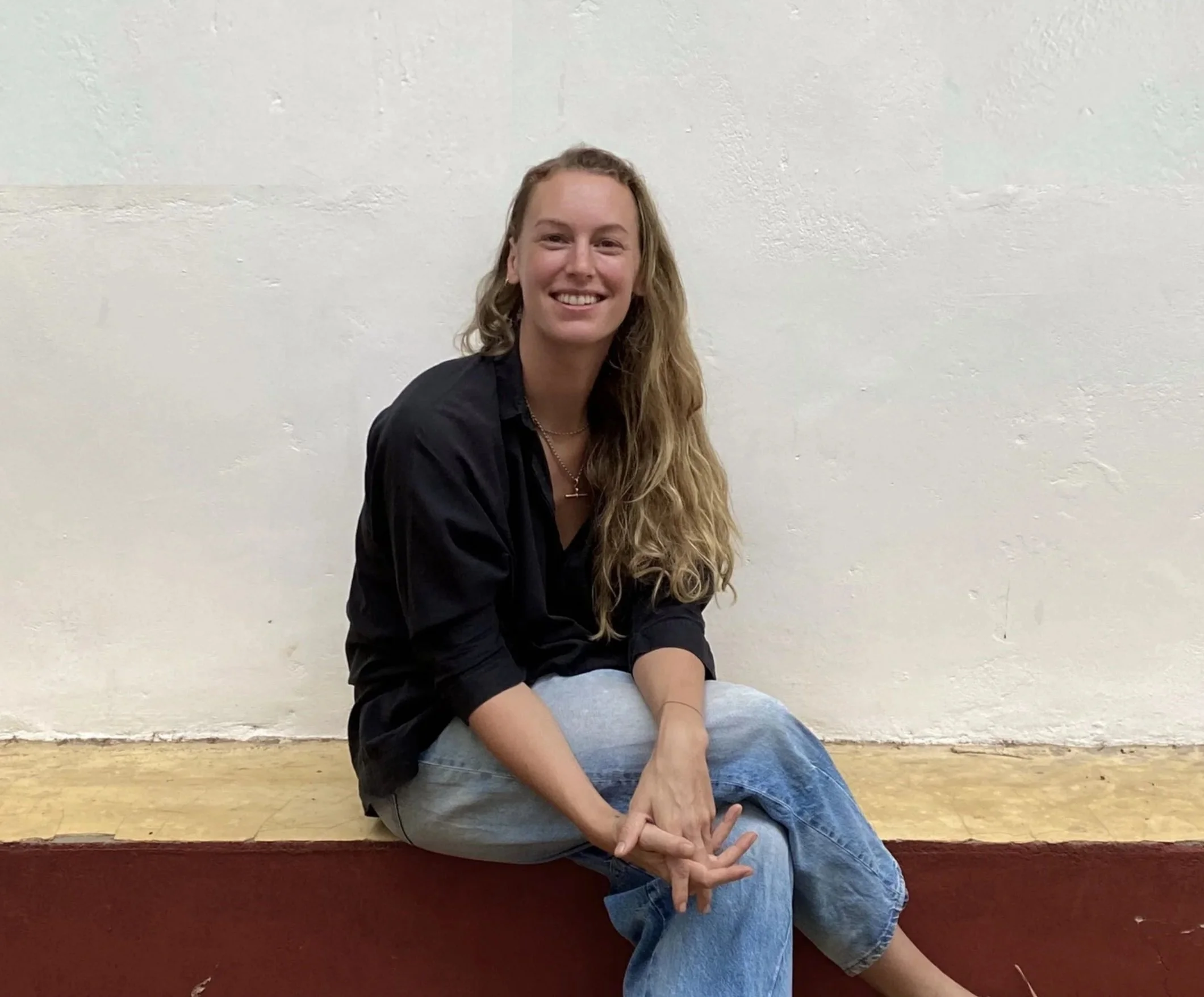 A young woman with long, curly blonde hair, sitting on a wooden bench against a plain white wall, smiling at the camera while wearing a black shirt and light-colored ripped jeans.
